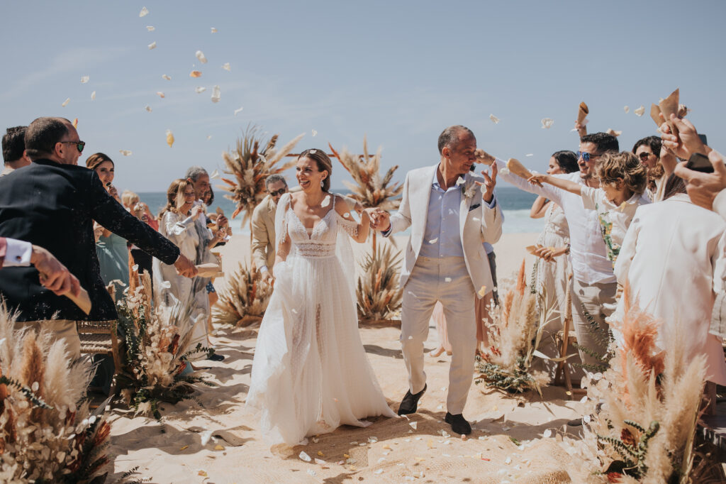 Bride and groom walk down sandy aisle, petals falling